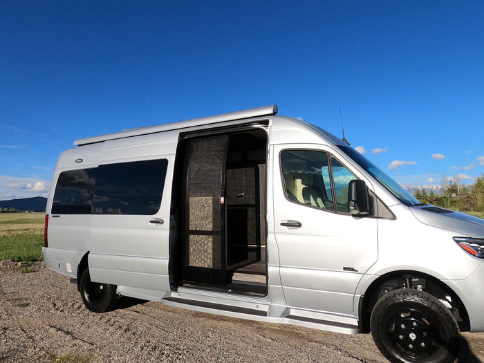 Silver RV rental camper van parked on gravel with its side door open, revealing a screen door, against a blue sky & mountains.