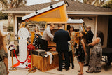 Guests enjoy a vintage orange RV rental camper van photo booth at an outdoor event, complete with fun props.