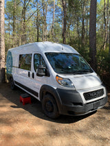 A white "Dave & Matt Vans" camper van (RV rental) is parked on a dirt path in a sunny forest, with a red step stool beside it.