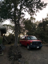 A red Ford van, resembling an RV rental, is parked among lush trees on a dirt path in nature.