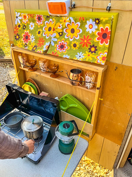 Cooking in a charming RV rental with retro floral curtains, vintage glasses, and a portable camping stove.