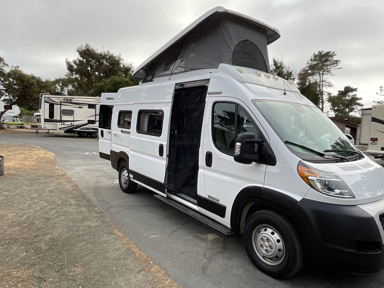 White pop-top camper van, an RV rental, parked at an RV park with its side door open under a cloudy sky.