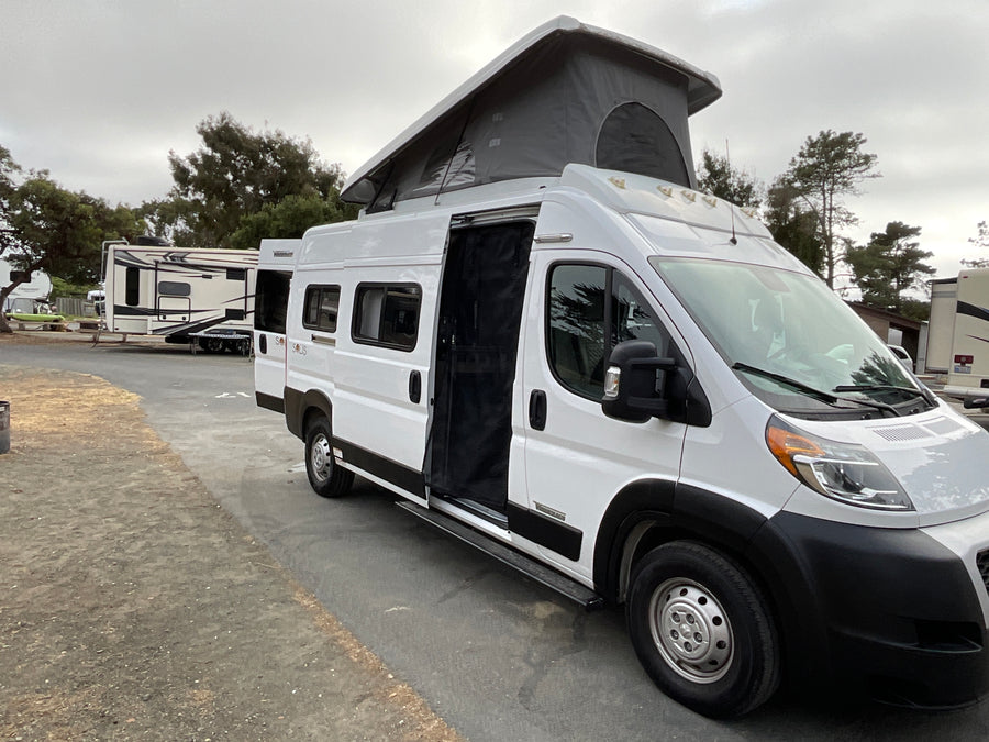 White pop-top camper van, an RV rental, parked at an RV park with its side door open under a cloudy sky.