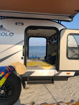 RV rental: Camper on sandy shore, open door reveals cozy bed, table, and stunning bridge view over water.