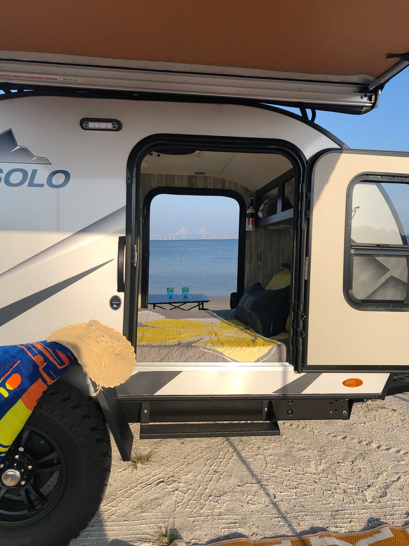 RV rental: Camper on sandy shore, open door reveals cozy bed, table, and stunning bridge view over water. — photo 8