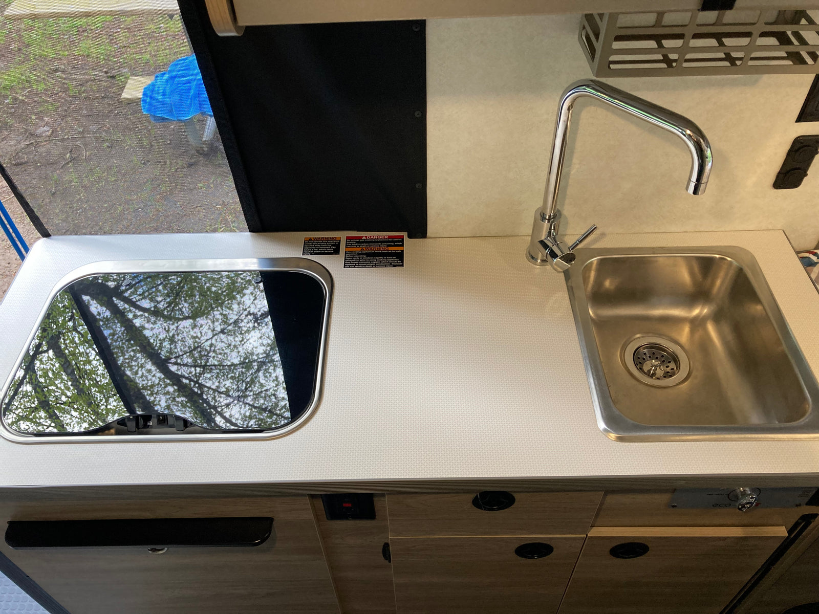 Compact rv rental kitchen with stainless steel sink, chrome faucet, and covered cooktop reflecting trees.