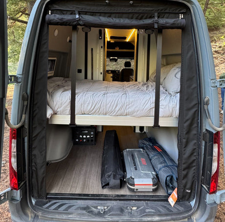 RV rental: Inside a camper van, showing a bed over under-bed storage on a wood floor, with open rear doors.