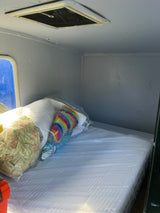 Clean RV rental sleeping area: new mattress in plastic, pillows, window, and ceiling vent in light interior.