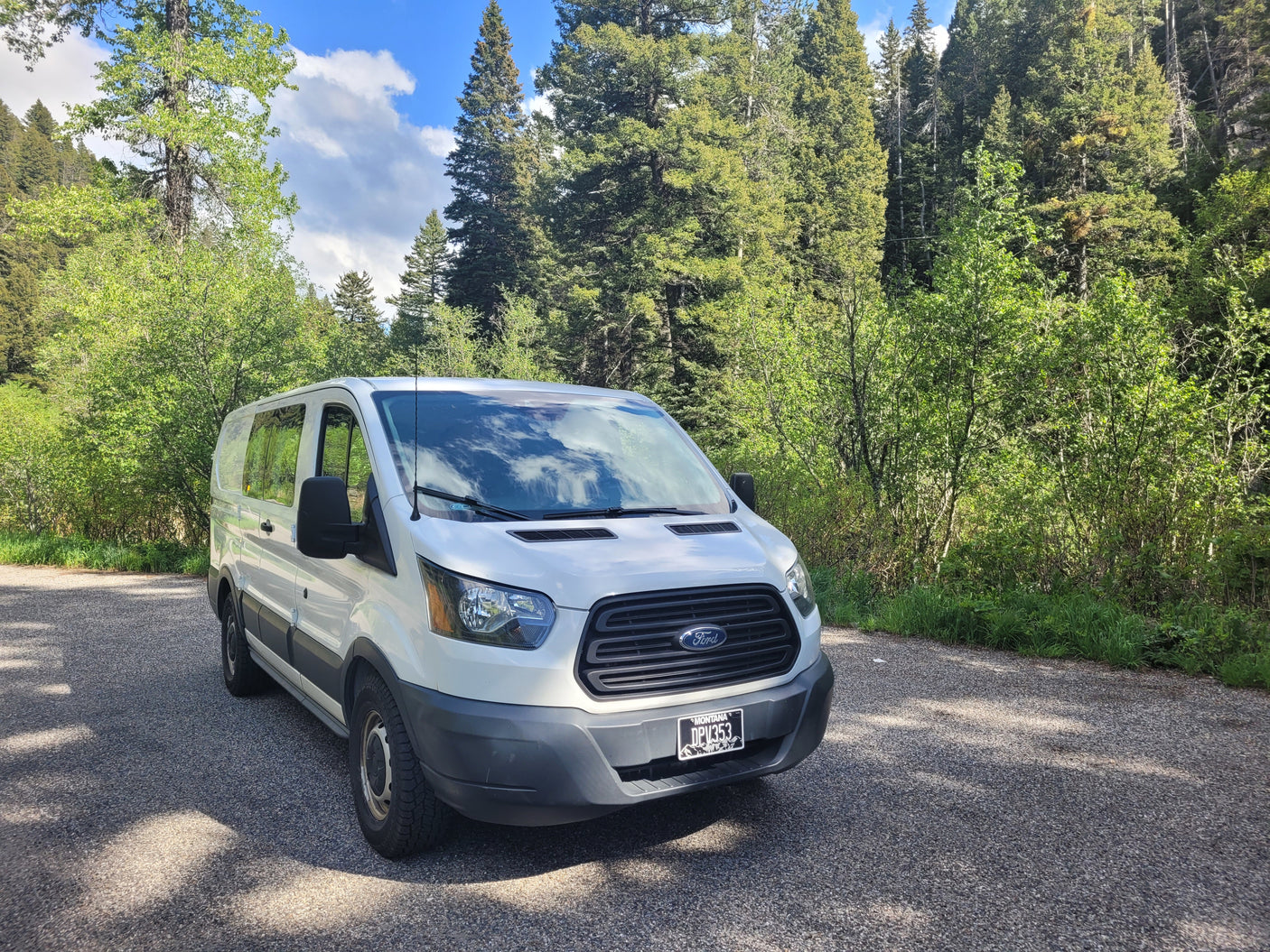 White Ford Transit camper van RV rental parked on an asphalt road, surrounded by lush green trees under a blue sky.