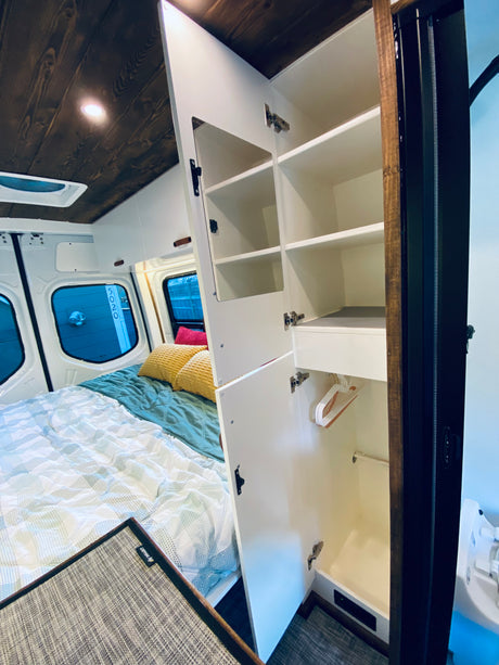 Spacious rv rental interior: comfortable bed, dark wood ceiling, and an open white wardrobe with shelves and hangers.