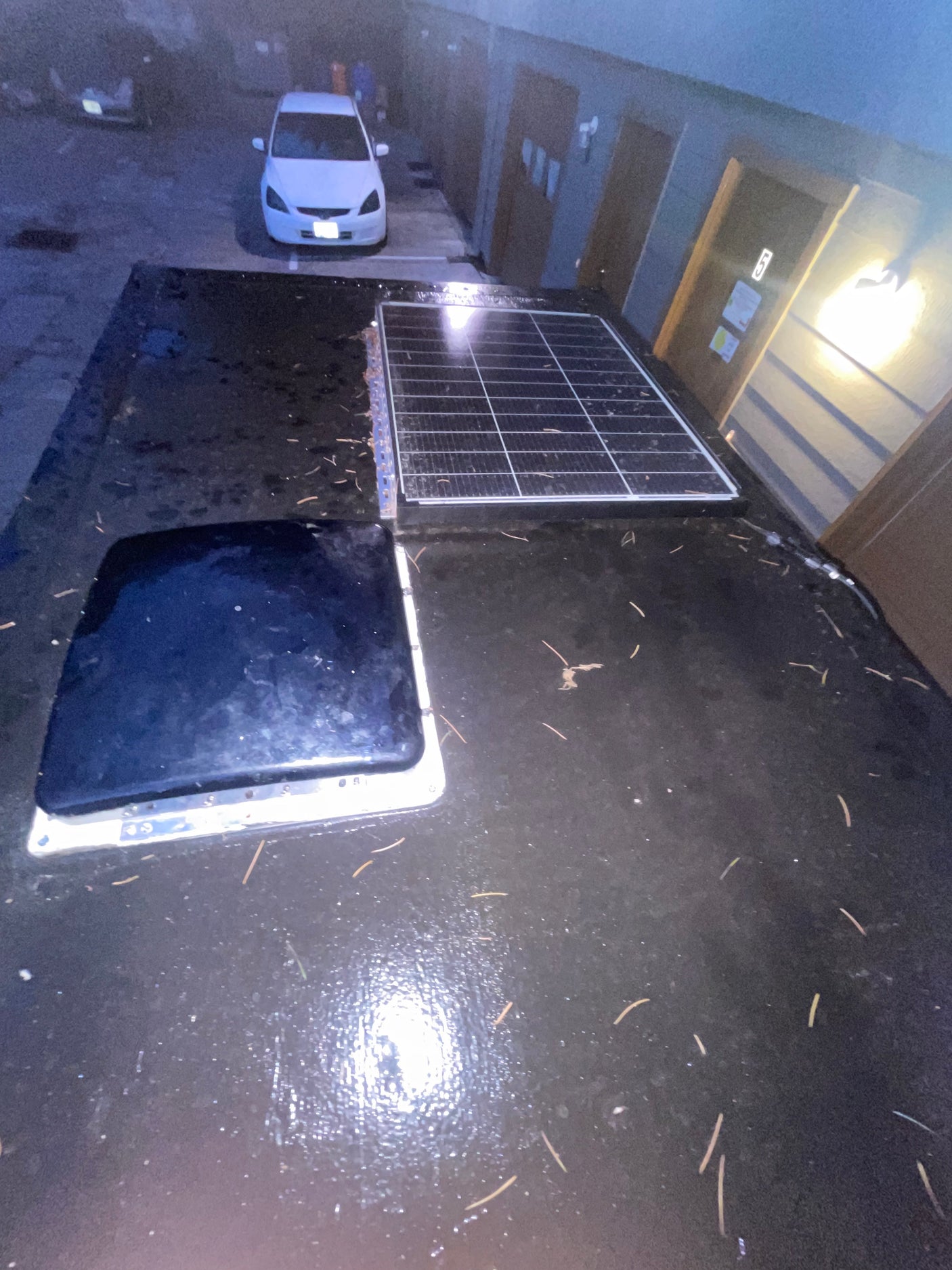 Overhead view of an RV rental's wet roof, featuring a solar panel and dark vent. Parked by buildings, ready for the road.