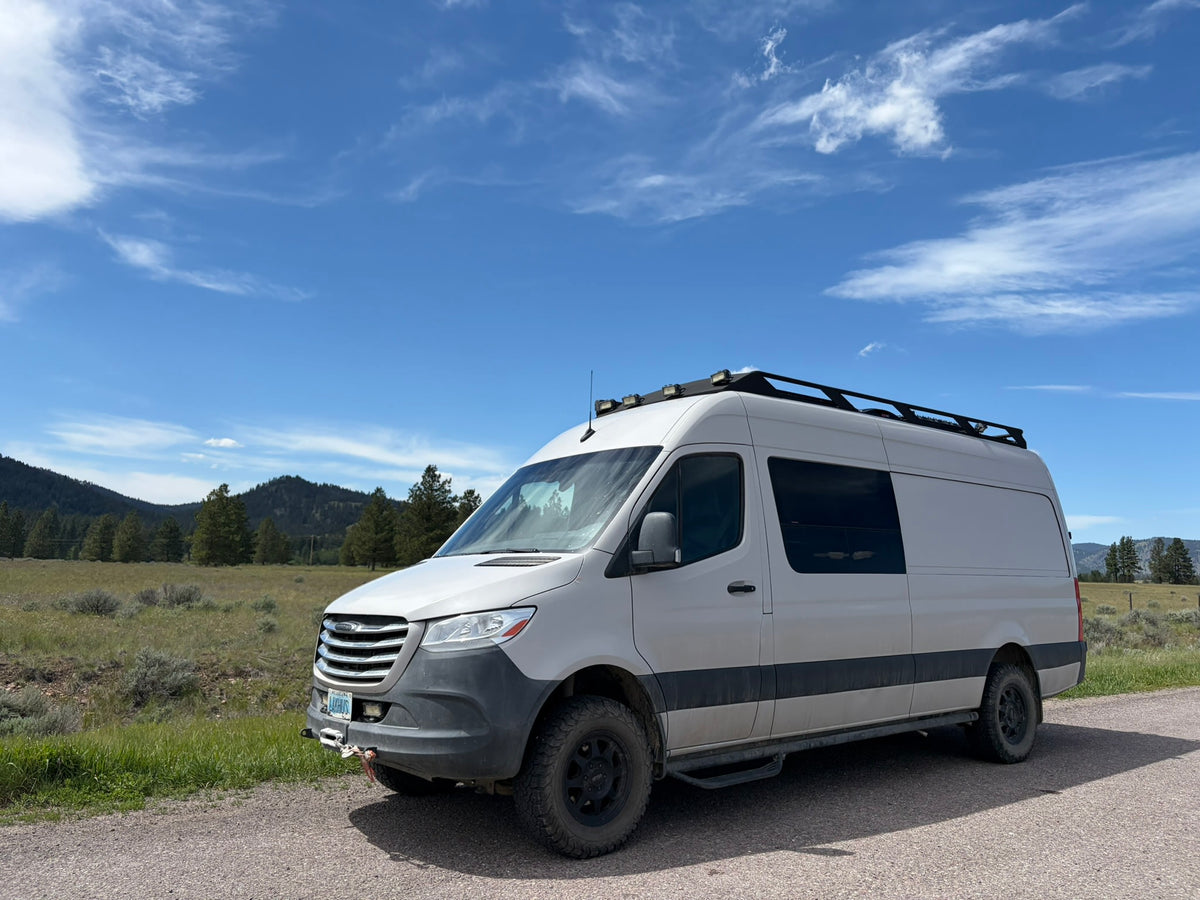 A white off-road camper van is parked on a gravel road with mountains and blue sky. Perfect for an rv rental adventure!|Experience van life with this RV rental: Features a raised bed