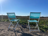 Two teal folding chairs and mugs facing a scenic field of green plants under a blue sky, ideal for an RV rental trip.