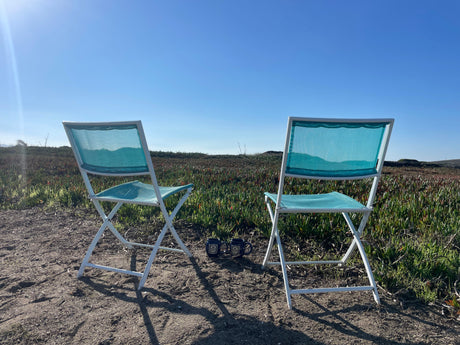 Two teal folding chairs and mugs facing a scenic field of green plants under a blue sky, ideal for an RV rental trip.