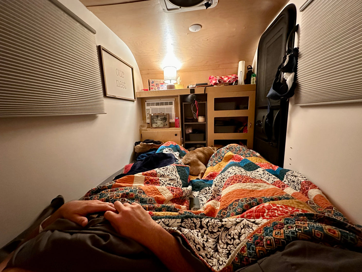 Cozy RV rental interior: Person and dog rest on comfortable bed under colorful quilt. Features compact storage and 'our nest' sign.