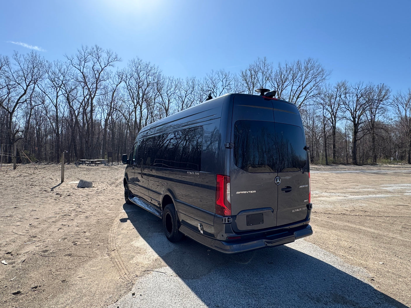 Dark gray Mercedes-Benz Sprinter luxury RV rental parked in a sandy lot by trees on a sunny day.