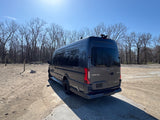 Dark gray Mercedes-Benz Sprinter luxury RV rental parked in a sandy lot by trees on a sunny day.