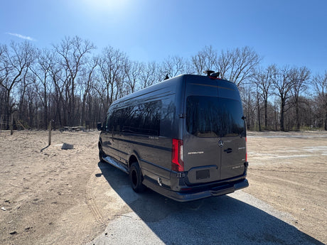 Dark gray Mercedes-Benz Sprinter luxury RV rental parked in a sandy lot by trees on a sunny day.