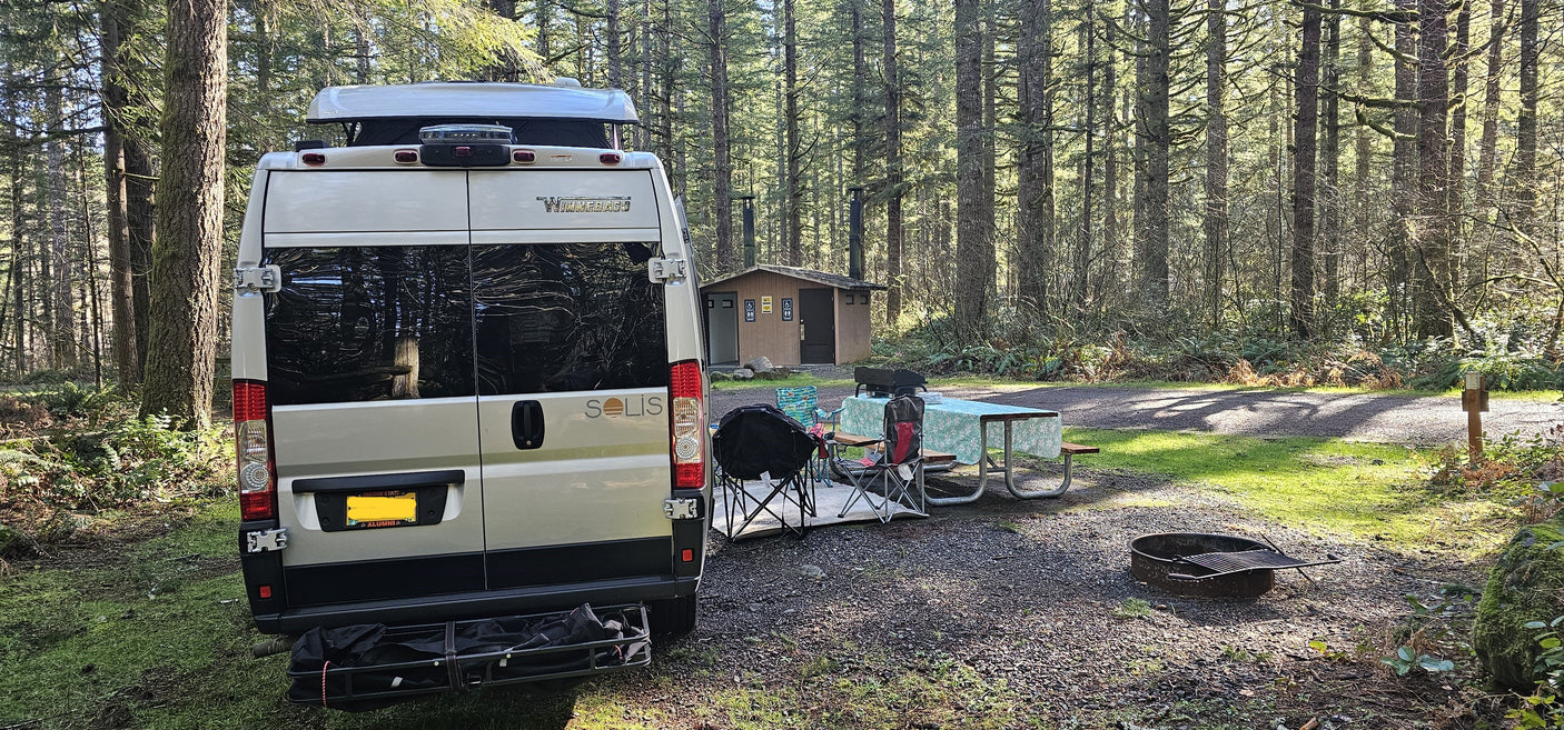 An RV rental camper van parked at a forested campsite with a picnic area, grill, and fire pit near restrooms.