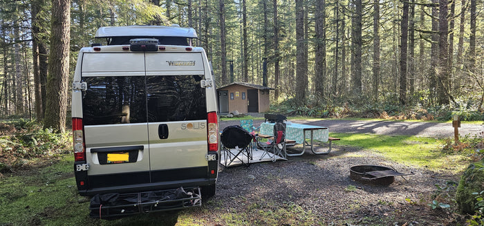 An RV rental camper van parked at a forested campsite with a picnic area, grill, and fire pit near restrooms.