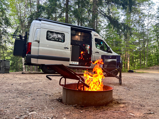 Enjoying an RV rental adventure! Camper van parked in a wooded campsite with a roaring fire pit & grill.