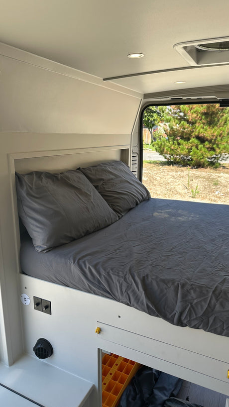 A comfortable bed with dark gray bedding and integrated charging ports inside a bright RV rental. Scenic outdoor view.