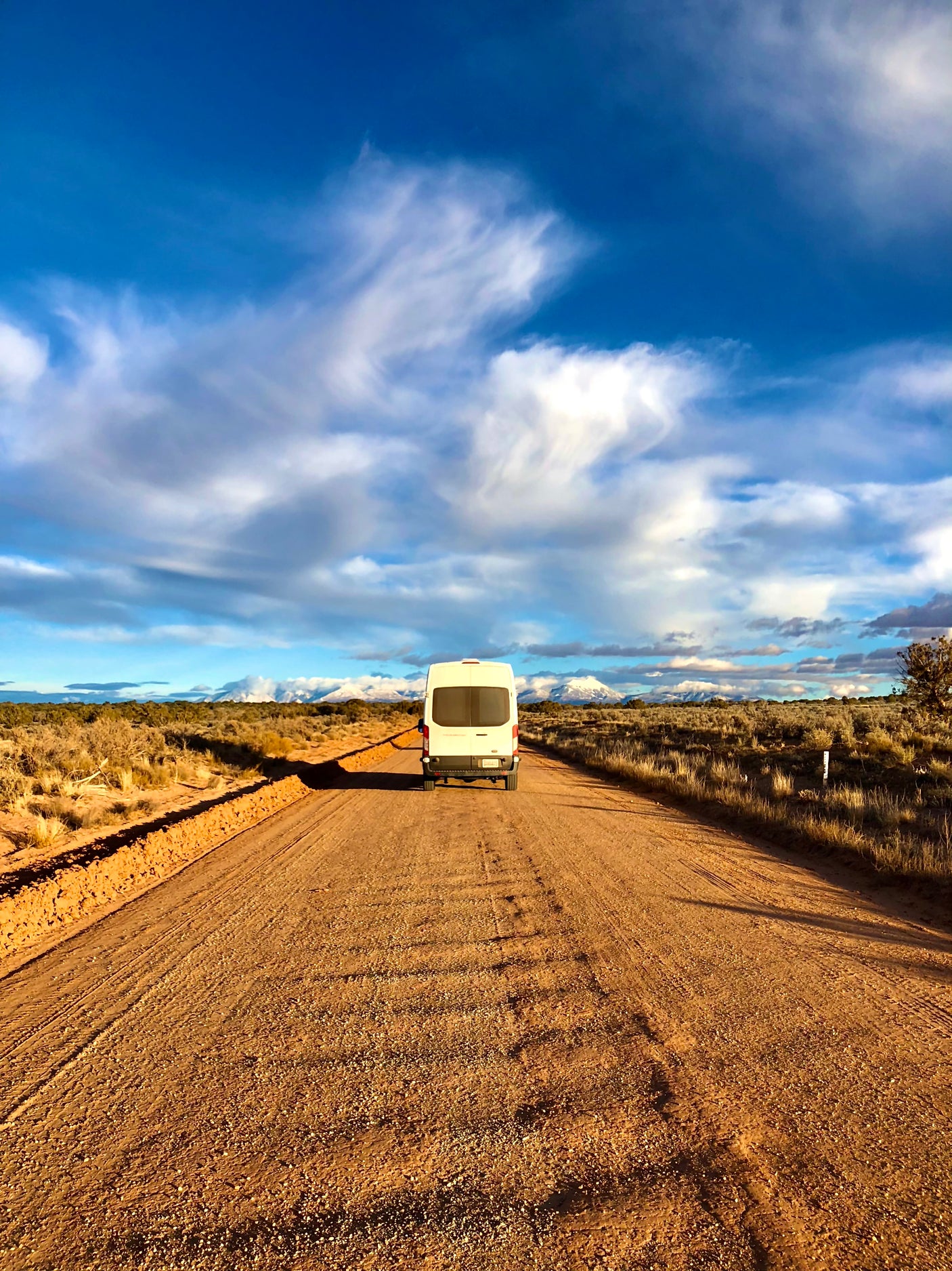 White RV rental camper van on a dusty desert road leading to distant snow-capped mountains under a dramatic sky.