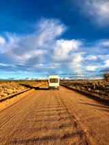 White RV rental camper van on a dusty desert road leading to distant snow-capped mountains under a dramatic sky.