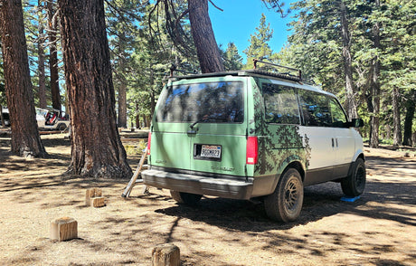 Customized green & white Astro van, an RV rental, sits in a sunny pine forest. Perfect for camping!