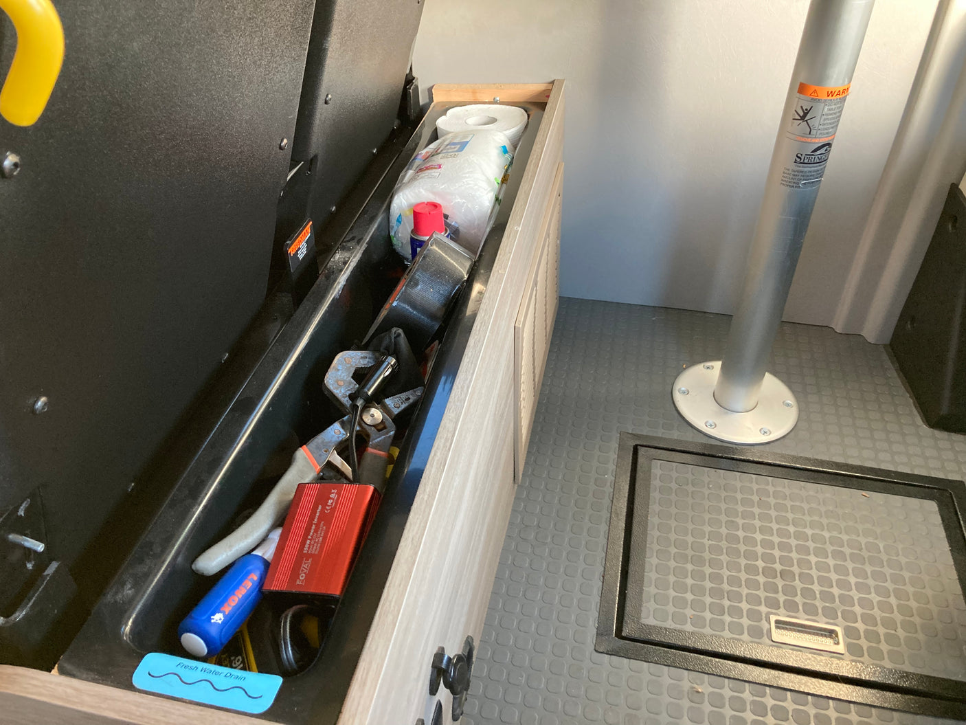 Deep storage in an rv rental with tools, a FOVAL power inverter, and paper towels. Gray floor, table leg, & floor hatch visible.