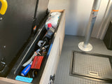 Deep storage in an rv rental with tools, a FOVAL power inverter, and paper towels. Gray floor, table leg, & floor hatch visible.