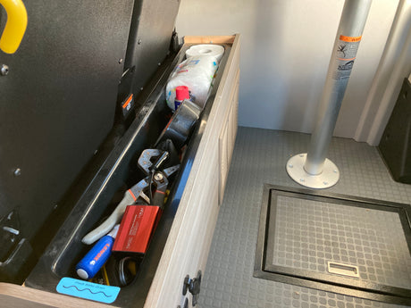 Deep storage in an rv rental with tools, a FOVAL power inverter, and paper towels. Gray floor, table leg, & floor hatch visible.