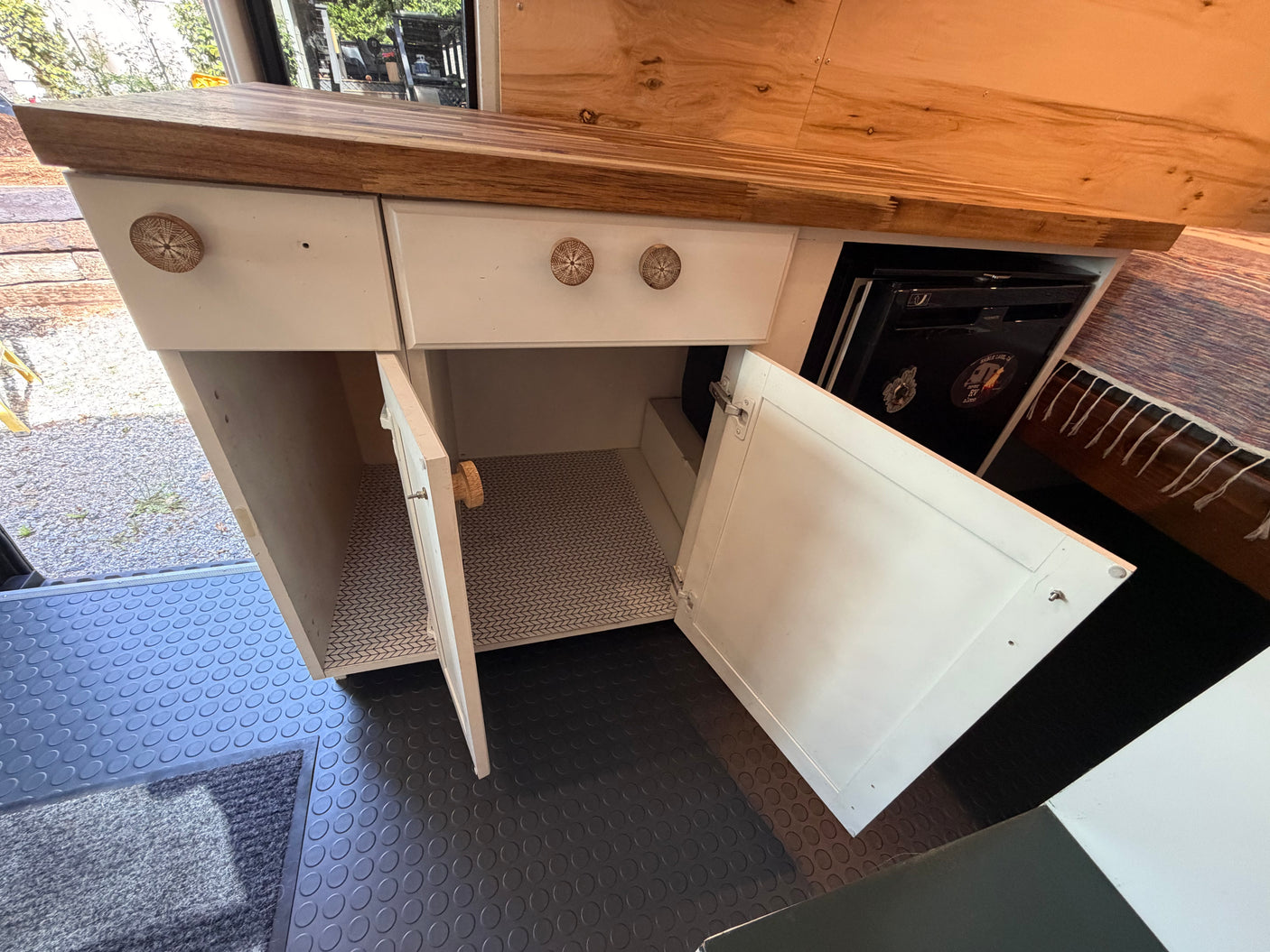 Detailed view of an RV rental camper van's kitchenette: white cabinets, wood counter, open storage, and built-in fridge.