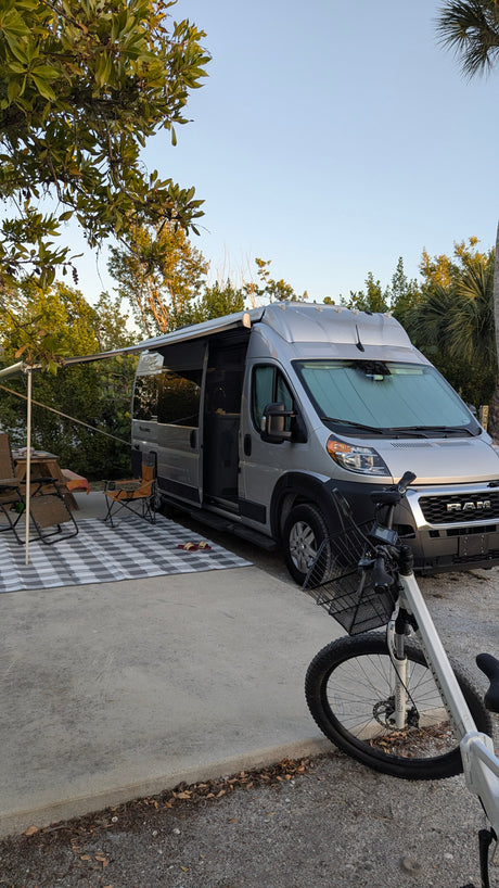 Silver camper van RV rental at campsite with extended awning, outdoor seating, and bike.