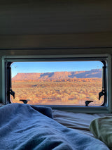 Cozy RV rental bed with a view of red rock desert mesas and scrubland through the window, offering a serene nature escape.