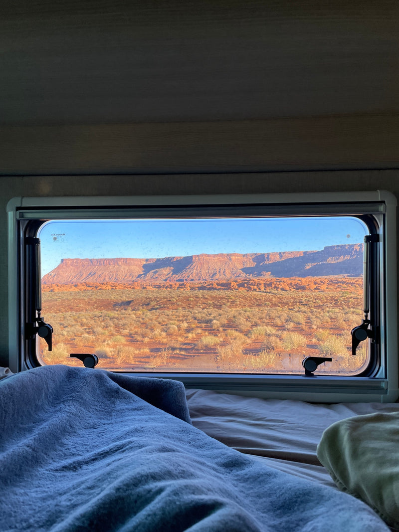 Cozy RV rental bed with a view of red rock desert mesas and scrubland through the window, offering a serene nature escape. — photo 14