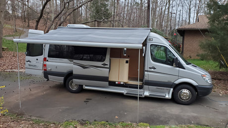 Silver Pleasure-Way Sprinter **rv rental** with extended awning & open door, parked on a driveway near trees.