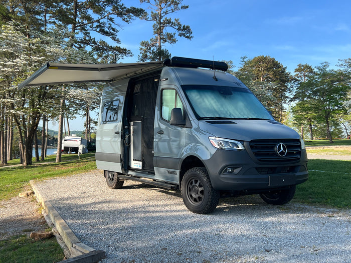 Modern gray Mercedes-Benz Sprinter RV rental camper van parked by a scenic lake with blooming trees, awning extended.