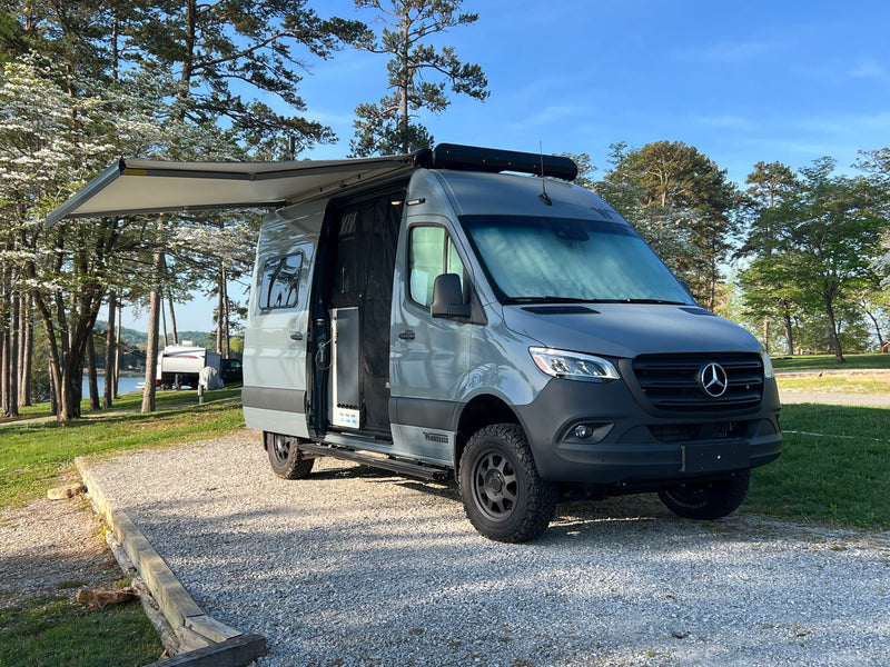 Modern gray Mercedes-Benz Sprinter RV rental camper van parked by a scenic lake with blooming trees, awning extended. — photo 3