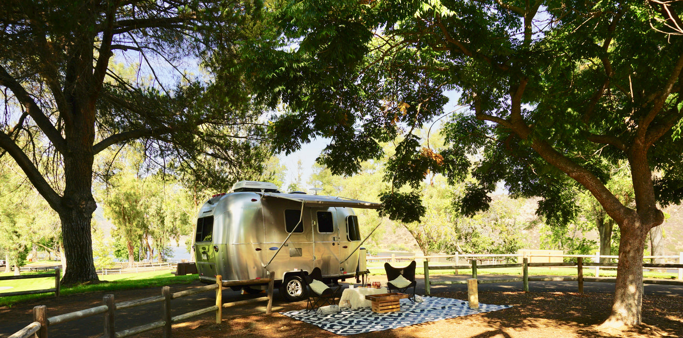 An Airstream RV rental is parked under trees, with an inviting outdoor lounge set up on a patterned rug by the water.
