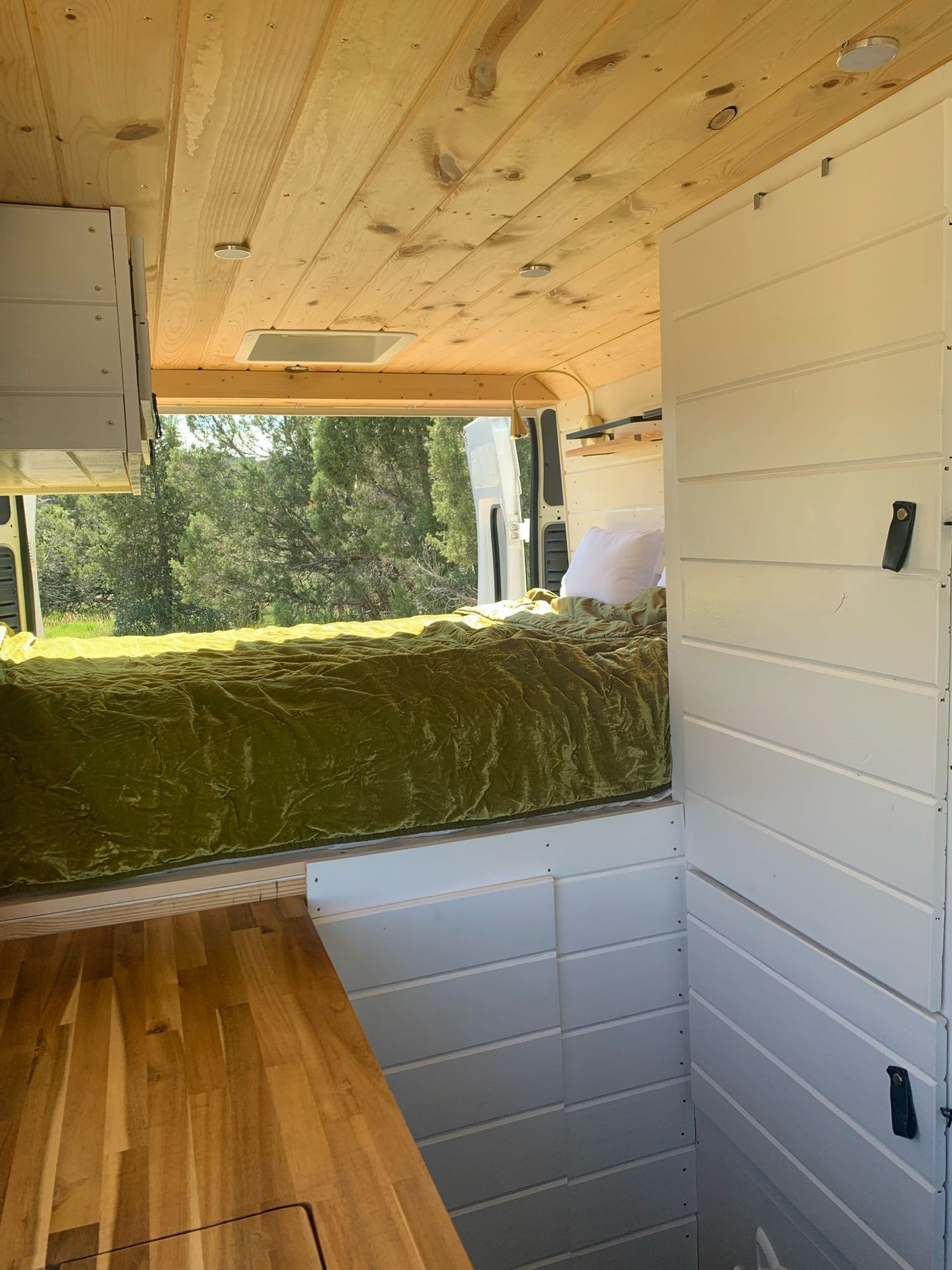 Inside this modern RV rental: a plush green bed, wood ceiling, white walls, and a stunning forest view. Ready for adventure.