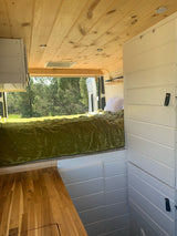 Inside this modern RV rental: a plush green bed, wood ceiling, white walls, and a stunning forest view. Ready for adventure.