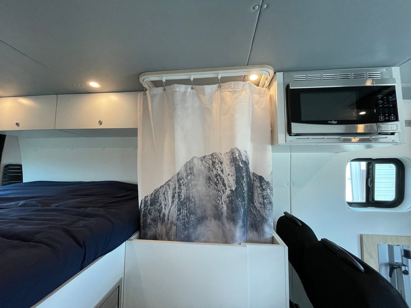 RV rental interior: a cozy bed, shower with a mountain graphic curtain, and overhead microwave. Compact, functional space. — photo 11