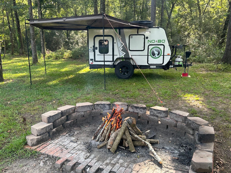 RV rental adventure! A white camper with an extended awning and a burning campfire in a brick pit, set in a natural, wooded area. — photo 1