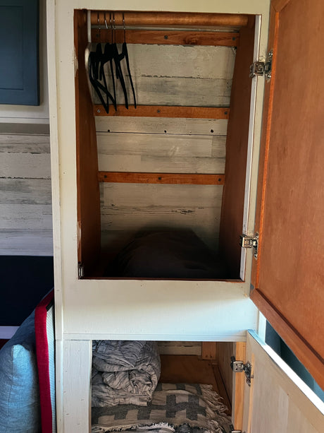 Inside an RV rental: a rustic closet with black hangers on a rod, wood plank walls, and neatly folded blankets below.