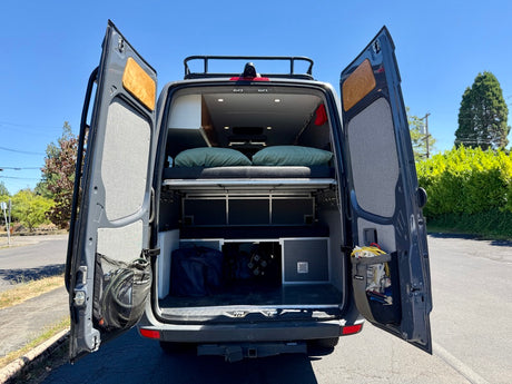 Spacious RV rental camper van with open rear doors reveals a cozy bed, ample storage, and organized gear for travel.