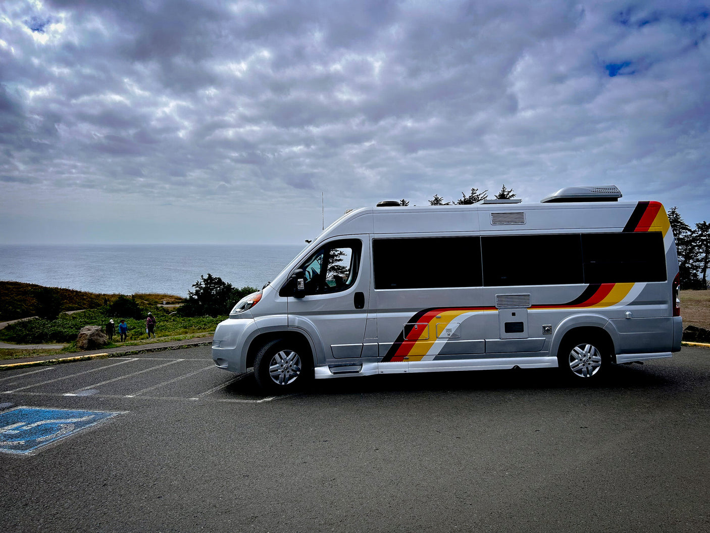 Silver RV rental with vibrant red, yellow, and black stripes, parked on a coastal overlook with the ocean and cloudy sky in the background.