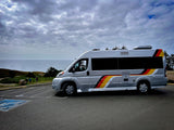 Silver RV rental with vibrant red, yellow, and black stripes, parked on a coastal overlook with the ocean and cloudy sky in the background.