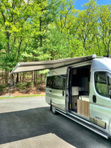An RV rental camper van is parked with its awning extended and door open, surrounded by lush green trees on a sunny day.