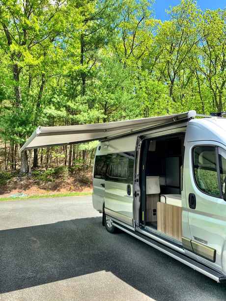 An RV rental camper van is parked with its awning extended and door open, surrounded by lush green trees on a sunny day.